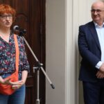 Kobieta z krótkimi rudymi włosami i okularami mówi do mikrofonu, ubrana w kwiecistą koszulę i pomarańczową torbę. Mężczyzna w garniturze stoi w pobliżu, uśmiecha się, a w tle widać obraz.