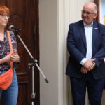 Kobieta z kręconymi rudymi włosami mówi do mikrofonu, podczas gdy mężczyzna w ciemnym garniturze i okularach stoi w pobliżu, uśmiechając się. Są w środku, obok obrazu i drewnianych drzwi.