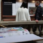 Notatnik i długopis leżą na biurku na pierwszym planie, a dwie osoby oglądają eksponaty muzealne na rozmytym tle. Uwaga skupiona jest na notatniku i długopisie.