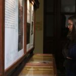 Dwie kobiety uważnie przyglądają się gablocie muzealnej i dużym ilustrowanym panelom informacyjnym zamontowanym na ścianie. Wydają się zamyślone i zainteresowane wystawą.