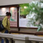 Kobieta z plecakiem i zieloną kurtką patrzy na wystawę z tekstem i obrazami na ścianie w pomieszczeniu. Na pierwszym planie widać rozmazane zielone i białe liście.