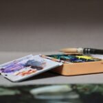 Otwarta paleta z farbami akwarelowymi w różnych kolorach i dwa pędzle na niej położone, ustawione na płaskiej powierzchni z rozmytym tłem.