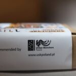 Zbliżenie małego pudełka tekturowego z białą etykietą, na której widnieje napis „Recommended by Urban Sketchers Poland” wraz z ich logo i stroną internetową www.uskpoland.pl. Pudełko jest lekko nieostre.