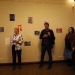 Grupa ludzi stoi w galerii sztuki, patrząc na małe dzieła sztuki wystawione na kremowej ścianie. Jedna osoba mówi, podczas gdy inna patrzy na telefon. Inni słuchają uważnie, niektórzy ze skrzyżowanymi ramionami.