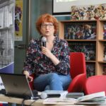 Kobieta z krótkimi rudymi włosami i okularami siedzi na czerwonym krześle, trzyma mikrofon i mówi. Ma laptopa na kolanach i komiksy na stole przed sobą. W tle widać regały z książkami i ekran.