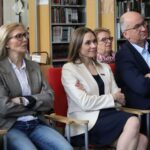 Grupa osób, zarówno mężczyzn, jak i kobiet, siedzi na czerwonych krzesłach ze skrzyżowanymi ramionami i uważnie obserwuje coś, co dzieje się poza ramką w pomieszczeniu z regałami i wystawionymi książkami.