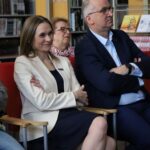 Trzech dorosłych siedzi w środku na czerwonych krzesłach, uważnie coś obserwując. Kobieta z przodu ma na sobie kremowy żakiet i ciemną sukienkę, lekko się uśmiecha, składając ręce. W tle widać regały z książkami.