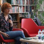 Kobieta z krótkimi rudymi włosami, okularami i wzorzystą bluzką siedzi na czerwonym krześle, trzymając mikrofon. Jest w bibliotece, za nią są regały z książkami. Na stole przed nią leżą dwie butelki wody i zeszyty.