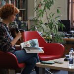 Kobieta z krótkimi rudymi włosami, w okularach i dżinsach, siedzi na czerwonym krześle, trzymając mikrofon i otwartą książkę. Znajduje się w bibliotece z regałami na książki, stołem i butelkami z wodą w pobliżu.