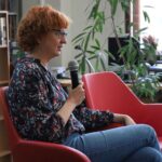 Kobieta z kręconymi rudymi włosami i okularami siedzi na czerwonym krześle, trzyma mikrofon i mówi. Ma na sobie kwiecistą bluzkę i dżinsy, a w tle widać regały z książkami i rośliny.