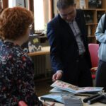 Mężczyzna w garniturze pokazuje otwartą ilustrowaną książkę siedzącej osobie z kręconymi włosami, ubranej w wzorzystą koszulę, wewnątrz pomieszczenia z regałami na książki, dużymi oknami i mikrofonami na stole.