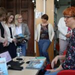 Grupa ludzi stoi i ogląda ilustrowane książki w bibliotece, podczas gdy kobieta o rudych włosach siedzi przy stole, przed nią leży więcej książek, stoi laptop i butelka wody.