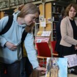 Młoda kobieta w okularach i jasnoniebieskiej bluzie z kapturem pochyla się nad stołem, aby przyjrzeć się obrazowi, podczas gdy inna kobieta stoi nieopodal w bibliotece, w tle widać regały z książkami i czerwone krzesła.
