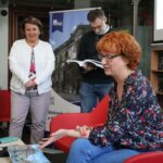 Czterech dorosłych znajduje się w bibliotece. Jedna kobieta z rudymi włosami siedzi i mówi, gestykulując ręką. Dwie kobiety i jeden mężczyzna stoją za nią, a w tle widać książki i baner.