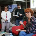 Czterech dorosłych znajduje się w bibliotece lub księgarni. Kobieta z krótkimi rudymi włosami rozmawia, siedząc na pierwszym planie; trzech innych stoi za nią, jeden czyta magazyn. Książki i plakaty wypełniają tło.