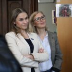 Dwie kobiety stojące w środku, jedna ze skrzyżowanymi ramionami w beżowej marynarce, a druga w okularach i kraciastej kurtce, obie patrzące przed siebie i lekko się uśmiechające. W tle widać regał z książkami i drzwi.