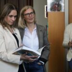 Trzy kobiety stoją w pomieszczeniu, każda z otwartą książką. Dwie kobiety patrzą przed siebie, jedna uśmiecha się delikatnie, podczas gdy trzecia kobieta po prawej czyta i pisze w swojej książce. W tle widać półki i plakaty.