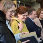 Pięciu dorosłych stoi blisko siebie w pomieszczeniu, a jedna kobieta na pierwszym planie przegląda książkę. Grupa wydaje się zaangażowana i uśmiechnięta, co sugeruje nieformalne spotkanie, prawdopodobnie w bibliotece lub biurze.