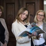 Trzy kobiety stoją w środku; jedna w środku, ubrana w jasną marynarkę, trzyma i patrzy na otwartą książkę Urban Sketching, podczas gdy dwie pozostałe kobiety patrzą i się uśmiechają. Drewniane drzwi są w tle.