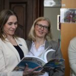 Trzy kobiety stoją razem w pomieszczeniu. Kobieta po lewej, ubrana w jasną marynarkę, uśmiecha się, patrząc na otwartą książkę, którą trzyma. Pozostałe dwie kobiety patrzą i się uśmiechają.