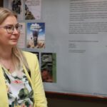Kobieta o długich blond włosach i okularach, ubrana w żółtą marynarkę nałożoną na kwiecistą bluzkę, stoi wewnątrz budynku przed tablicą z tekstem i obrazami.