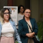 Trzy kobiety stoją w pomieszczeniu, dwie z przodu i jedna lekko za nimi. Kobiety z przodu noszą okulary i neutralne wyrazy twarzy; jedna nosi kurtkę dżinsową, druga turkusową marynarkę. W tle widoczny jest plakat wydarzenia naukowego.