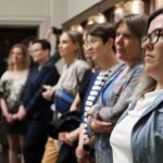 Grupa ludzi, głównie kobiet, stoi w środku w linii, uważnie patrząc w tym samym kierunku. Wydaje się, że słuchają kogoś lub czegoś poza kadrem. Sceneria wygląda jak wystawa lub formalne wydarzenie.