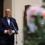 Mężczyzna w niebieskim garniturze mówi do mikrofonu w pomieszczeniu, stoi przy drewnianych drzwiach i tablicy informacyjnej, na pierwszym planie widać rozmazane kwiaty.