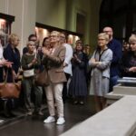 Grupa ludzi stoi w środku, niektórzy klaszczą, a inni uważnie się przyglądają. Są ubrani w stroje casualowe lub biznesowe, a na ścianie za nimi widać tablice informacyjne. Na pierwszym planie znajduje się duża roślina doniczkowa.