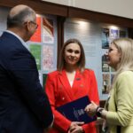 Trzy osoby stoją w pomieszczeniu i rozmawiają. Kobieta w czerwonej marynarce trzyma teczkę i się uśmiecha, stojąc twarzą do dwóch innych osób — mężczyzny w niebieskim garniturze i kobiety w jasnej marynarce — na tle tablic informacyjnych.