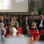 Pięć osób siedzi w panelu przed publicznością na wydarzeniu książkowym w bibliotece. Za nimi są regały z książkami, rośliny i ekran prezentacyjny z polskim tekstem. Jeden z panelistów trzyma mikrofon i przemawia.