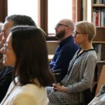 Grupa ludzi siedzi w środku, uważnie słuchając czegoś poza zasięgiem wzroku. W tle znajdują się regały z książkami i okna, a większość osób patrzy w prawo.