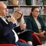 Mężczyzna mówi do mikrofonu, a dwie kobiety uważnie słuchają obok niego; wszystkie trzy siedzą na czerwonych krzesłach przed regałami z książkami.