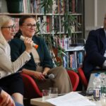Trzy osoby siedzą na krzesłach podczas dyskusji panelowej w bibliotece. Jedna kobieta mówi do mikrofonu, podczas gdy inni słuchają i się uśmiechają. Na stole z przodu znajdują się regały z książkami, roślina i butelki z wodą.