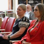 Dwie kobiety siedzą uważnie na czerwonych krzesłach podczas wydarzenia w pomieszczeniu. Jedna ma na sobie czarny wzorzysty top, a druga czerwoną kurtkę. W tle widać mężczyznę i regały z książkami.