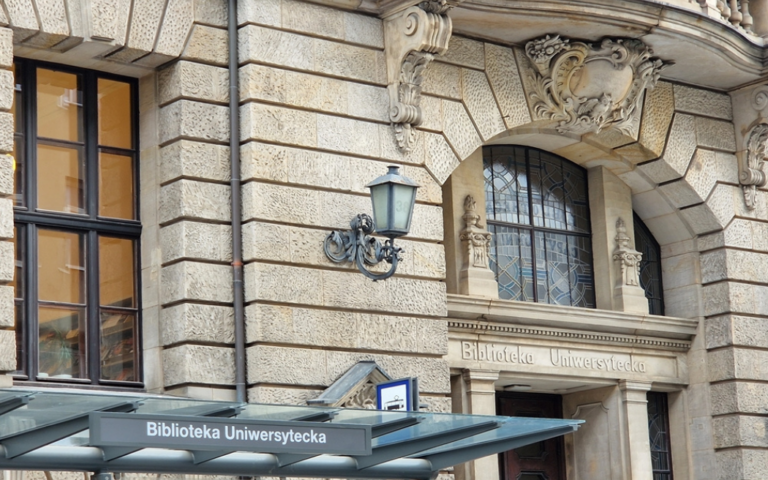 Zabytkowy kamienny budynek z łukowymi oknami i ozdobnymi detalami architektonicznymi. Nad wejściem i na zadaszeniu na pierwszym planie widoczny jest napis Biblioteka Uniwersytecka.
