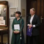 Dwie kobiety stoją obok siebie przy mikrofonach w pomieszczeniu, trzymając teczki. Jedna z nich ma na sobie zieloną sukienkę, a druga czarną marynarkę. Za nimi znajdują się plakaty informacyjne i drewniane drzwi.