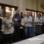 Grupa ludzi stoi uważnie w muzeum lub galerii, obserwując eksponaty wyświetlane na ścianach, z panelami informacyjnymi i oprawionymi dokumentami za nimi.