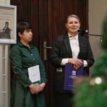 Dwie kobiety stoją w pomieszczeniu przy drewnianych drzwiach, jedna w zielonej sukience trzyma teczkę, druga w ciemnym garniturze mówi do mikrofonu i trzyma niebieską teczkę. Obok nich widoczny jest oprawiony plakat i broszury.