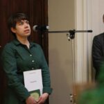 Kobieta w zielonej sukience stoi przy mikrofonie trzymając certyfikat, podczas gdy inna kobieta w szarej marynarce przygląda się i uśmiecha w tle. Znajdują się w pomieszczeniu w pobliżu drewnianych drzwi i udekorowanej choinki.