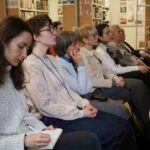 Grupa kobiet siedzi w rzędach krzeseł na zamkniętym wydarzeniu. Niektóre uważnie słuchają, podczas gdy jedna kobieta na pierwszym planie robi notatki w notatniku. W tle widoczne są półki z książkami.