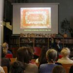 Grupa osób siedzi w bibliotece przed ekranem wyświetlającym prezentację o Danielu Fischerze w Wchows, 1754-1757, z obrazem czerwonej tkaniny. W tle widoczne są półki z książkami i rośliny.