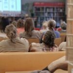 Ludzie siedzący na ławkach w bibliotece lub klasie, zwróceni w stronę ekranu, na którym wyświetlane są niewyraźne obrazy lub slajdy. Scena sugeruje publiczność uczestniczącą w prezentacji lub wykładzie.