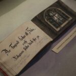 Stara, otwarta książka z ozdobnym niemieckim pismem na jednej stronie i ilustracją okna na drugiej, wyświetlana na szarej powierzchni obok małej etykiety.