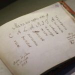 Stara, otwarta książka zawiera odręcznie zapisane łacińskie słowa ułożone ukośnie i pionowo, wraz z notatkami i podpisami wykonanymi brązowym atramentem; papier wydaje się postarzały i lekko pożółkły.