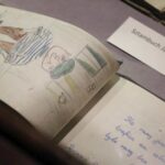 Zbliżenie otwartego notatnika przedstawiającego rysunek kolorowym ołówkiem osoby w pasiastej koszuli i kapeluszu. Obok znajduje się odręcznie zapisana strona i etykieta z polskim tekstem.