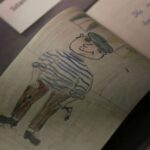 Dziecięcy rysunek uśmiechniętej osoby ubranej w pasiastą koszulę, brązowe spodnie i czapkę, trzymającej w jednej ręce jakiś przedmiot. Rysunek znajduje się wewnątrz otwartego zeszytu wystawionego za szybą.