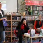 Kobieta stoi i przemawia, trzymając w ręku broszurę w bibliotece. Dwie inne kobiety siedzą przy stole z książkami i papierami, słuchając. W tle widoczne są półki z książkami i rośliny.