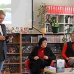 Trzy kobiety znajdują się w bibliotece; jedna stoi czytając gazetę, podczas gdy dwie siedzą obok stołu z wystawionymi książkami. W tle znajdują się półki z książkami i roślina, a na ścianie częściowo widoczny jest wyświetlany obraz.