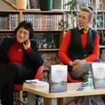 Dwie kobiety siedzą na czerwonych krzesłach przy stole z książkami, wodą i papierami. Znajdują się w bibliotece lub księgarni, z półkami kolorowych książek za nimi. Obie kobiety noszą okulary i wydają się być zaangażowane w rozmowę lub dyskusję.